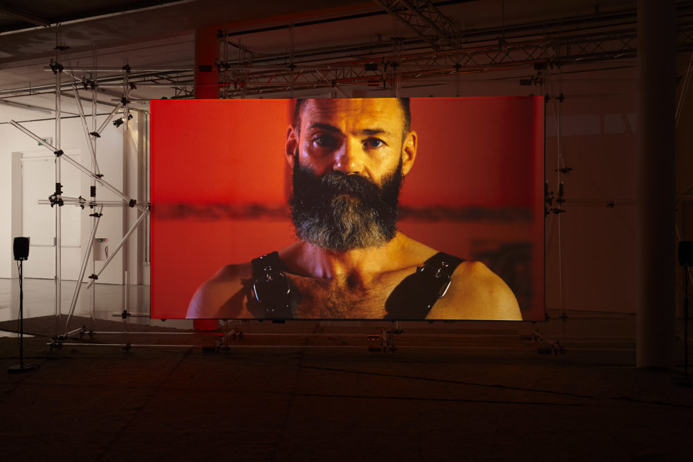 A screen has been erected at Spike Island gallery using a lot of scaffolding. On the screen, a person with a full dark beard is wearing leather bondage straps and looking directly at the camera.