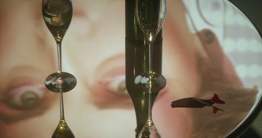Film still: Wine glasses and a bottle sit on a mirrored table, alongside a fortune telling fish that would come from a cracker.