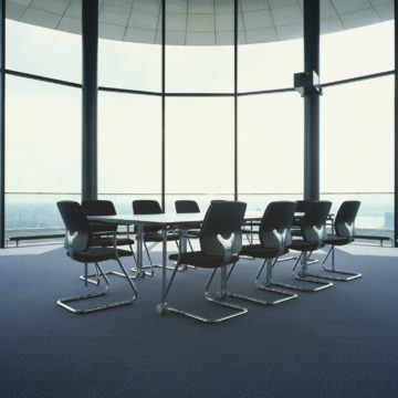 A table set up for board meetings sits in an otherwise empty room with walls made of glass. It seems this room is the tallest for miles.