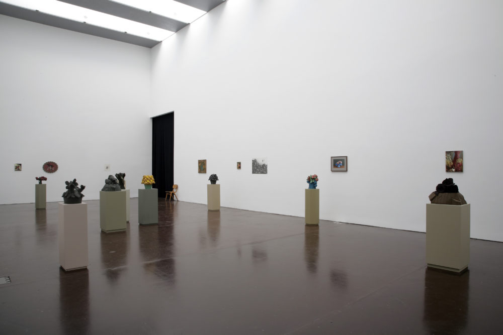 Installation view of Prophet (2008). Geometric sculptures made of clay sit on plinths. There are eight plinths in total. and a collection of framed floral images on the walls of the gallery.