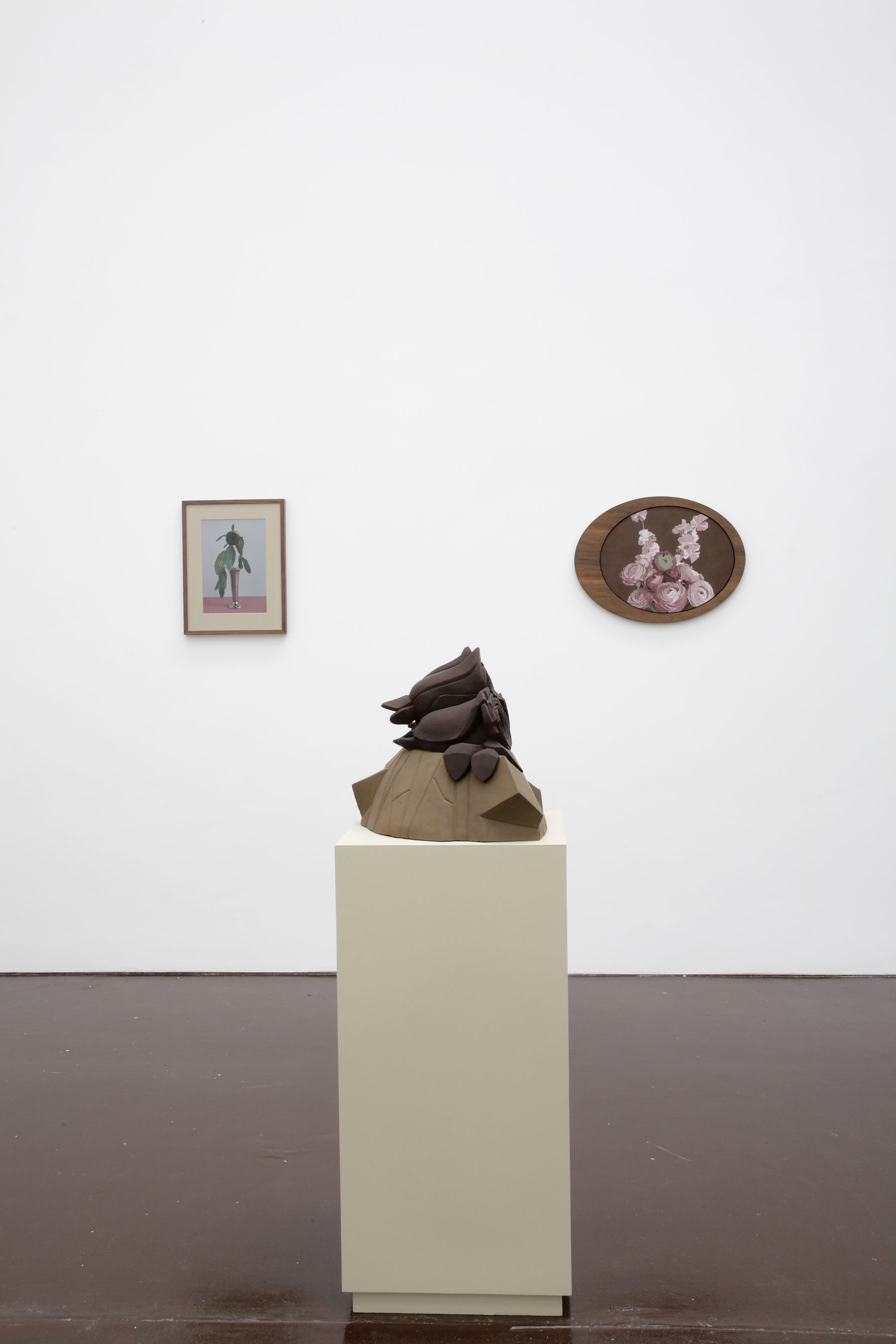 Installation view of Prophet (2008). A geometric sculpture made of clay sits on a plinth. Two floral images hang in frames behind.