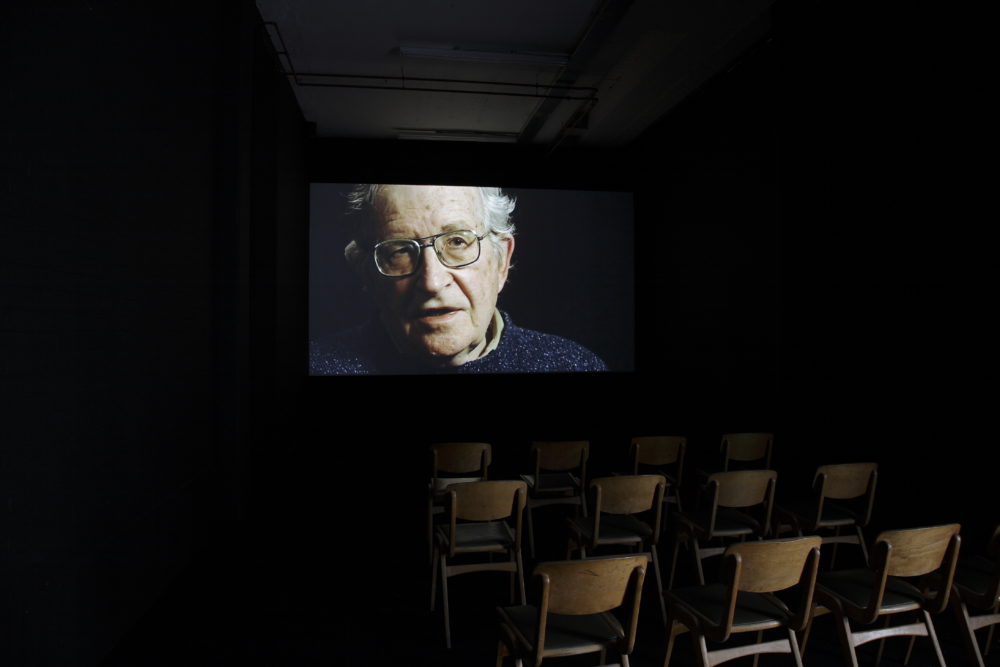Installation view of Prophet (2008). Wooden chairs sit in rows in the dark facing a projection of an elderly man.