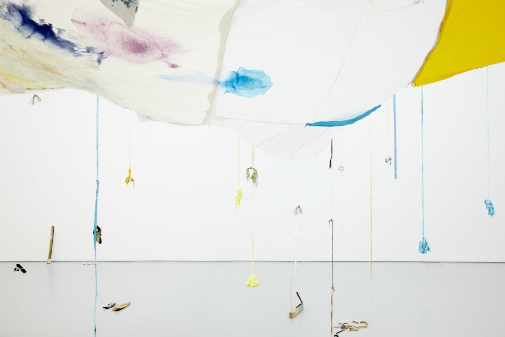 Large swathes of fabric hang down from the gallery ceiling. A white piece of fabric has been discolored in patches with pink and blue inks. Ribbons hang from these fabric swathes, some have weights which appear to be abstract shapes made from ceramic.