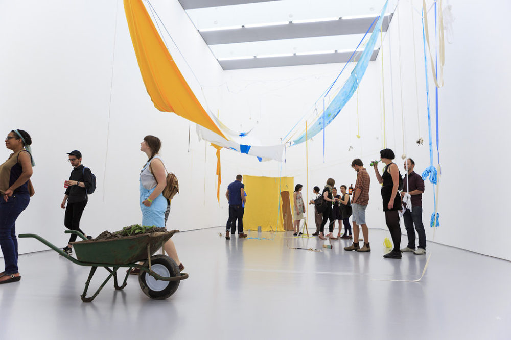 Visitors study the art in the gallery - in the foreground a wheelbarrow full of mud or clay. In the background a swathe of sunny yellow fabric is hung on a wall. The ceiling has more fabric hanging from it - this time in yellow, blue and white.