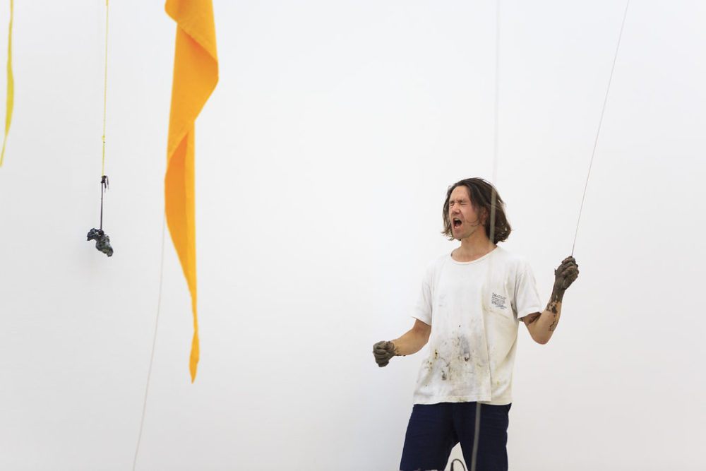 Yellow fabric hangs from the ceiling in a bright white gallery space. A man stands in a white t-shirt with hands covered in clay and some clay on his shirt. He has his eyes closed and he is shouting while pulling on some string. To the left, a fired clay abstract structure hangs on a piece of string.