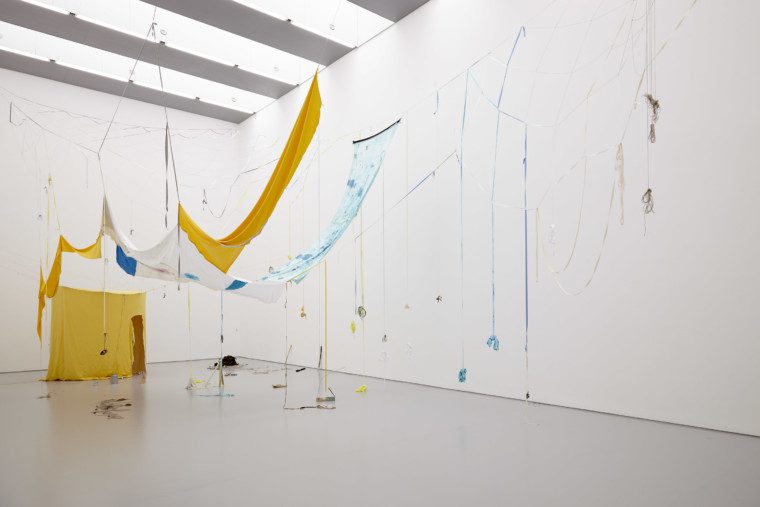 Photograph of a large white gallery space with large windows across the ceiling. A yellow tent-like structure is hanging in the distance, and swathes of fabric hang down from the ceiling in yellow and blues. Ribbons dangle from the fabric strips, and some have small clay objects tied to them.