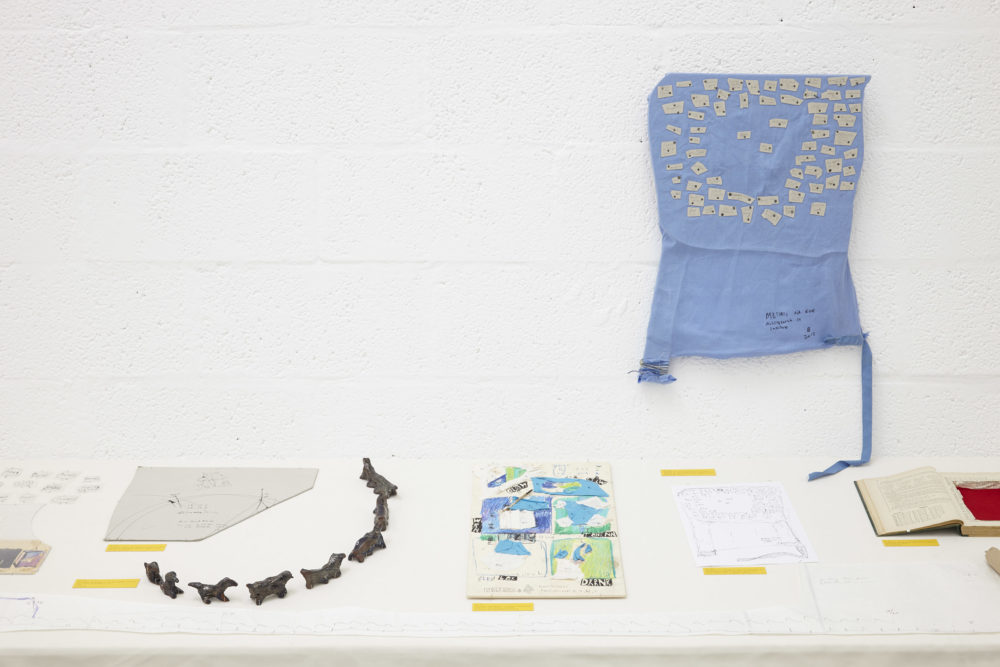 A white table in a white gallery has various ephemera laid out neatly and labelled.