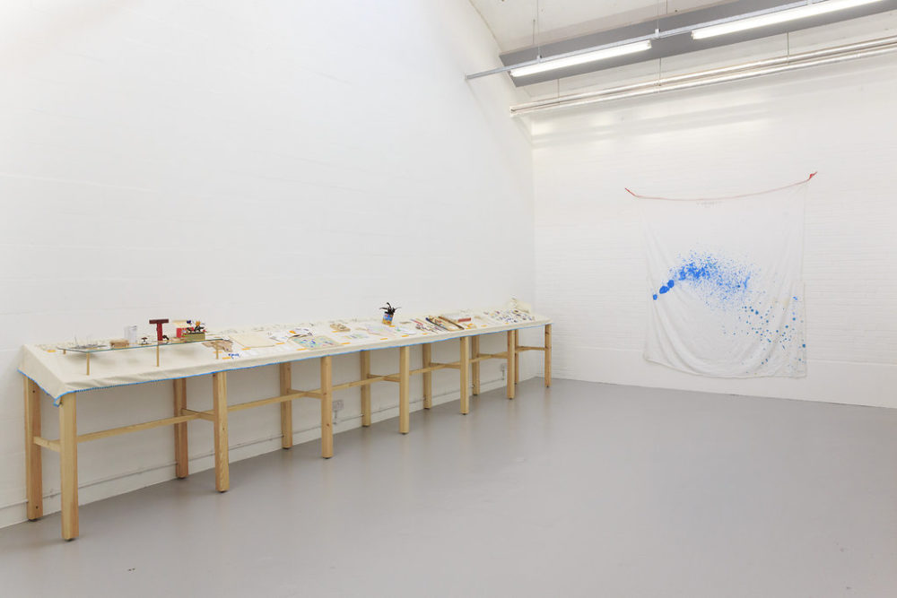 Various ephemeral pieces sit on a table in the white gallery. On another wall a piece of white fabric is hung up with paint sprayed on it.