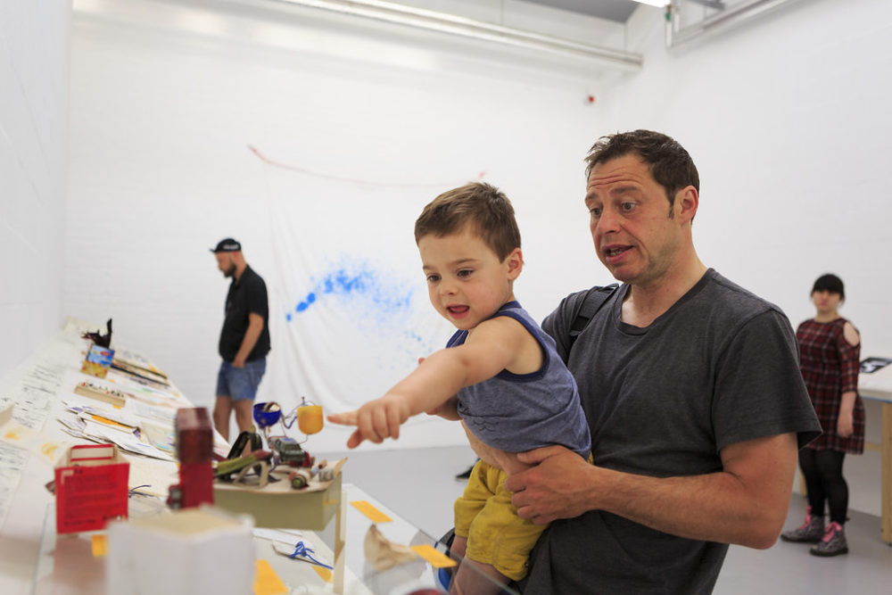 Two visitors of different ages (a toddler and a grown-up) point at various ephemeral pieces on a table in the white gallery. The toddler seems to have found a kinder egg cup.