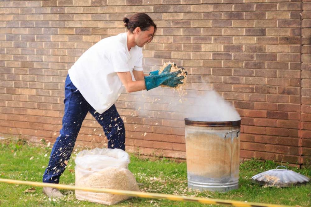 The artist is putting sawdust into a burning metal dustbin in order to make a rudimentary kiln. This is happening outside of the Spike Island building - some yellow and black tape prevents visitors from getting too close.