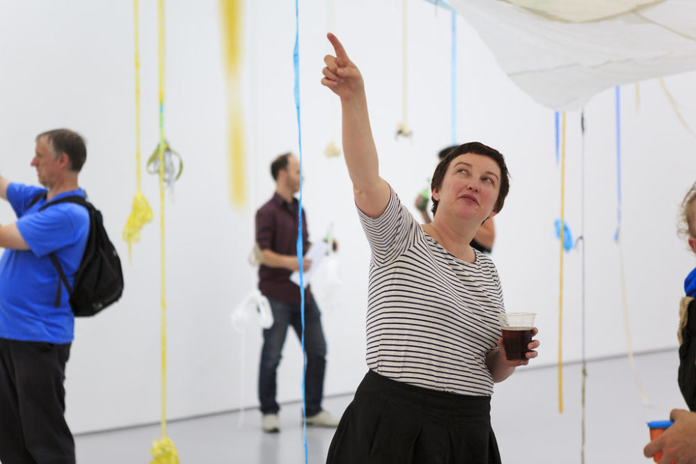 A visitor wearing a striped shirt points upwards. Around her, ribbons of blue, yellow and white hang down from the ceiling.