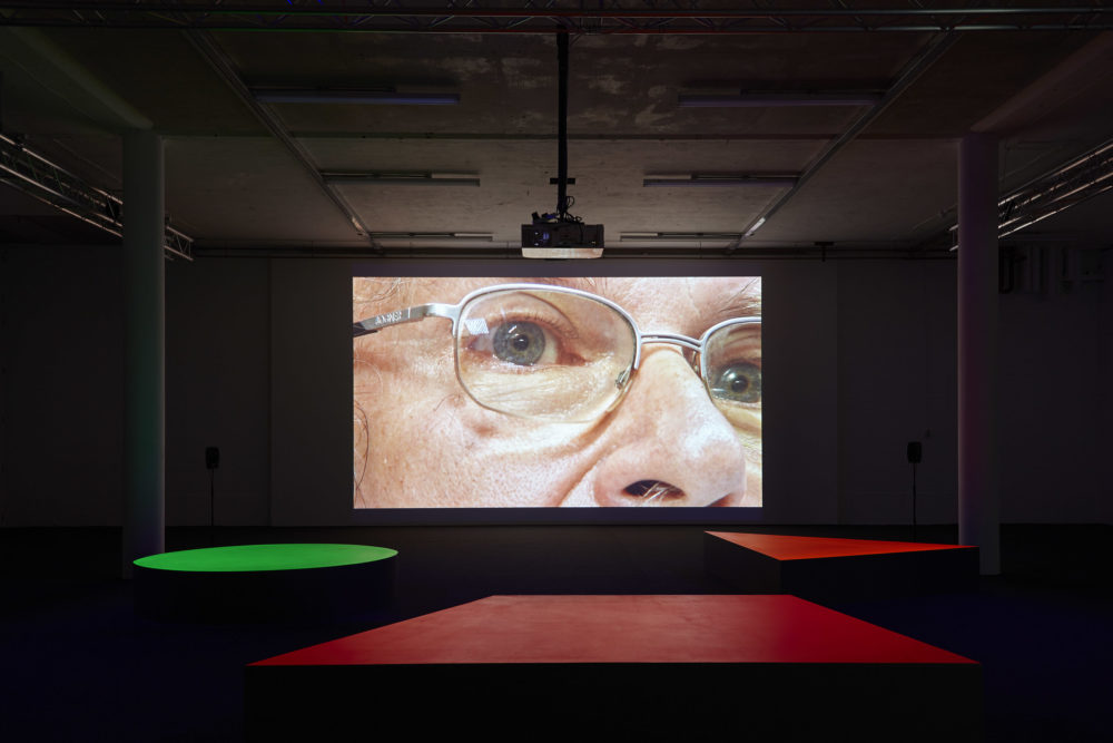 A projection of a man's eyes and nose very close up. The floor in the gallery is black with large brightly coloured geometric benches in front of the screen.