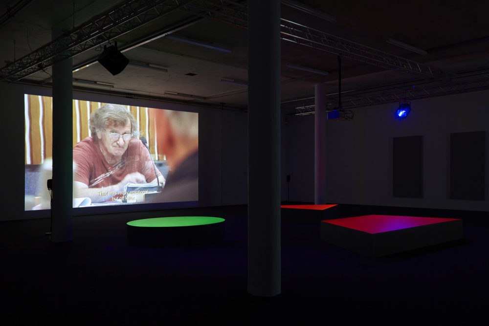 A projection of a man looking at the camera in deep concentration. The floor in the gallery is black with large brightly coloured geometric benches in front of the screen.