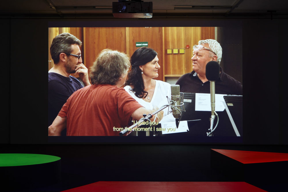 Three men and a woman stand in a recording studio. Subtitles read 