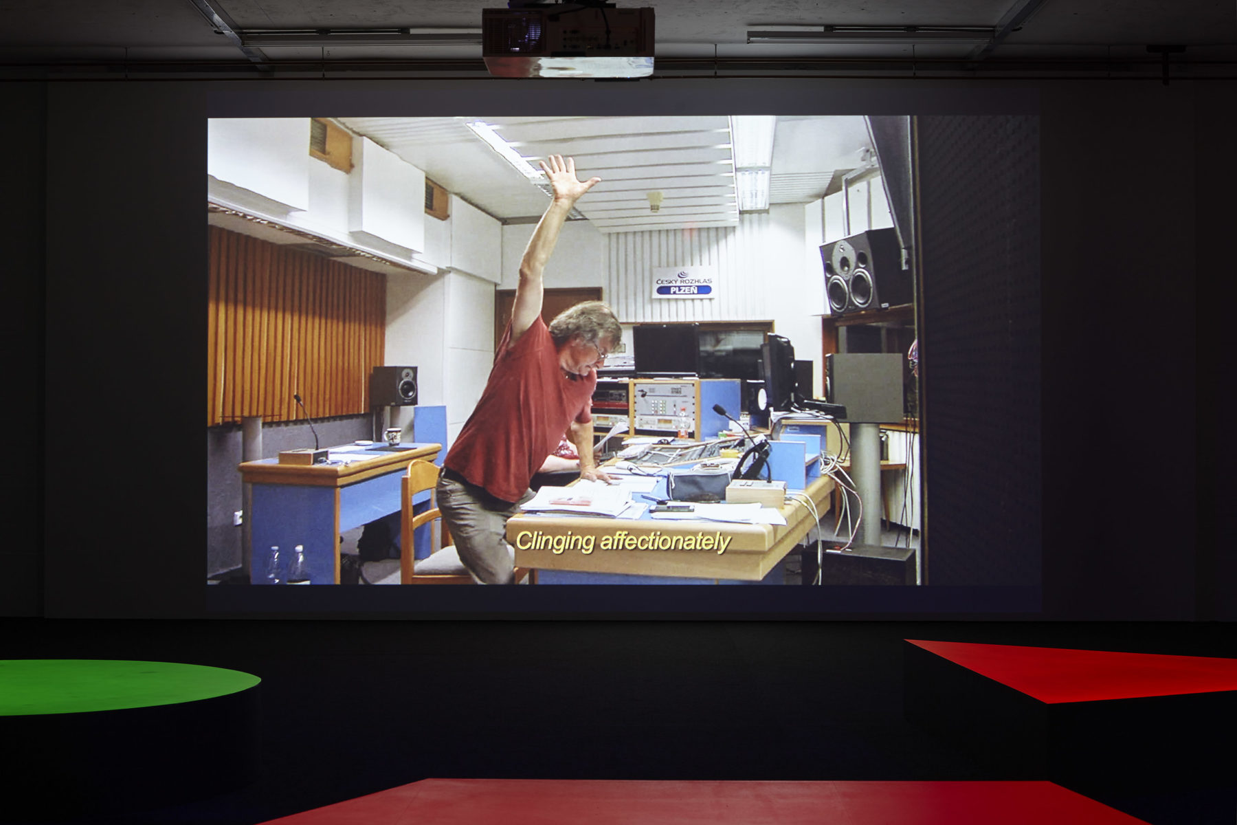 A projection of a man stood over a table full of papers and electronic equipment, holding one arm upwards. Subtitles read 