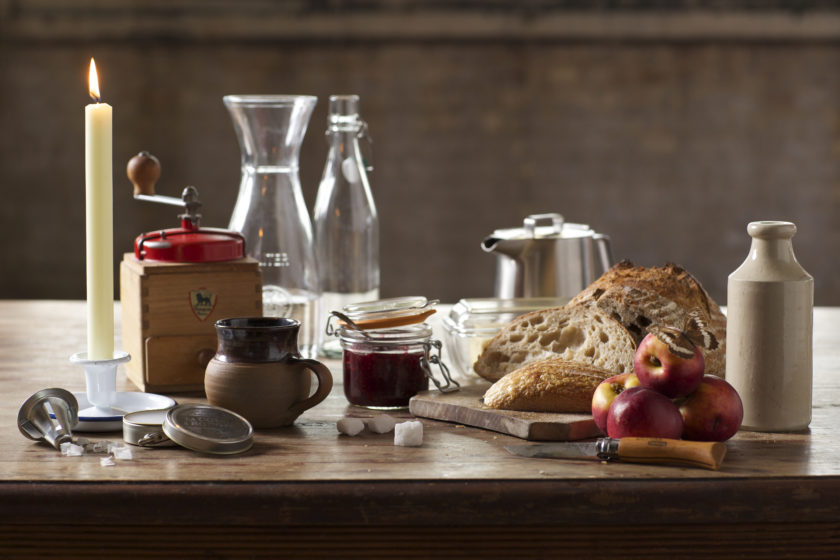 A table scene - a coffee grinder, a pot of jam, a loaf of bread, some apples all sit on a table.