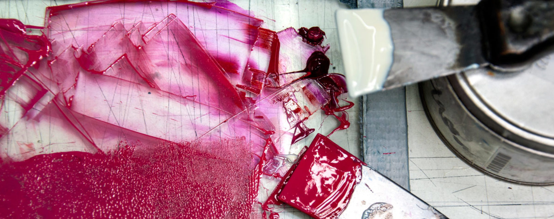 A close up of paint scrapers with glossy pink and white paint on them. The paint has been scraped onto a perspex surface.