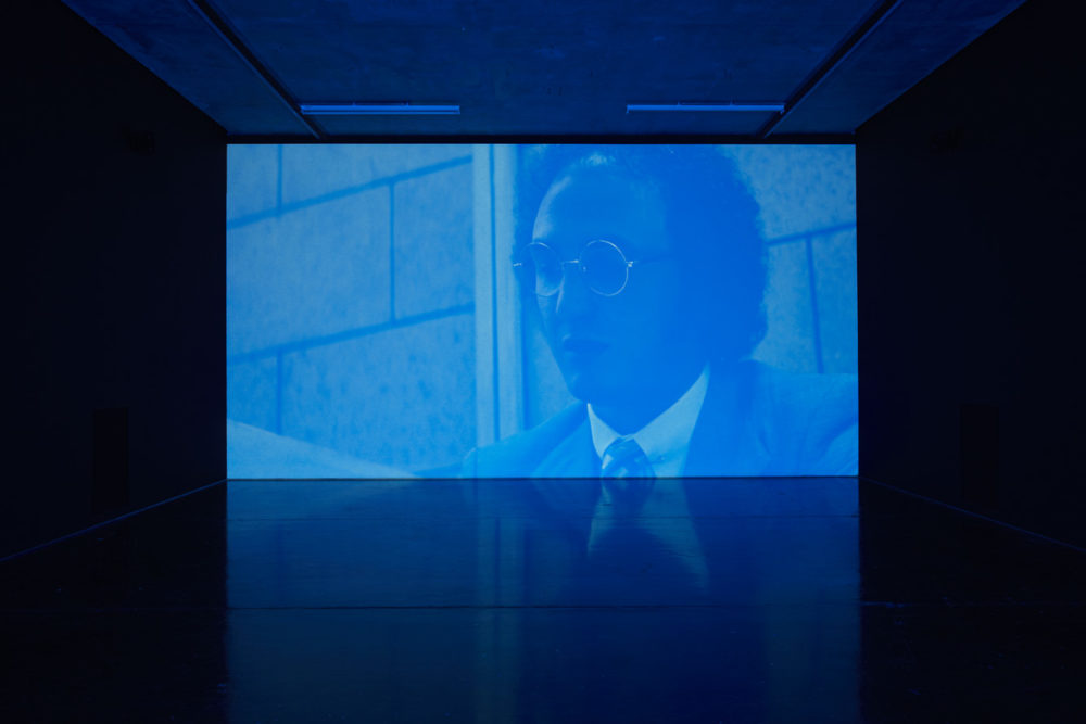 Installation view of Savage, I Asked You a Thousand Times (2011). A blue hued projection of a man in a suit.