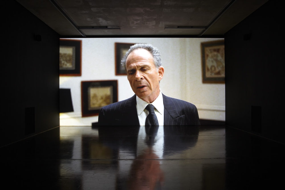 SInstallation view of Savage Presents Jean Michel Jarre (2011). A projection of a beleaguered looking man with grey hair in a suit.