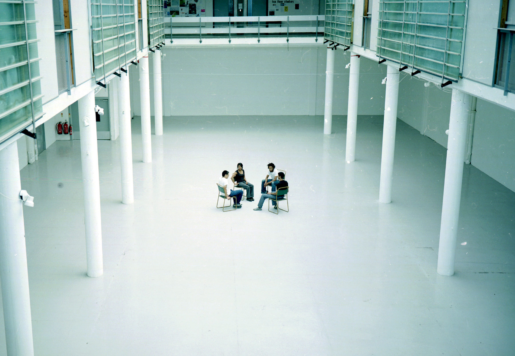 Installation view of Spin Cycle (2004). Four people sit on chairs facing one another in the centre of a white, bright gallery.