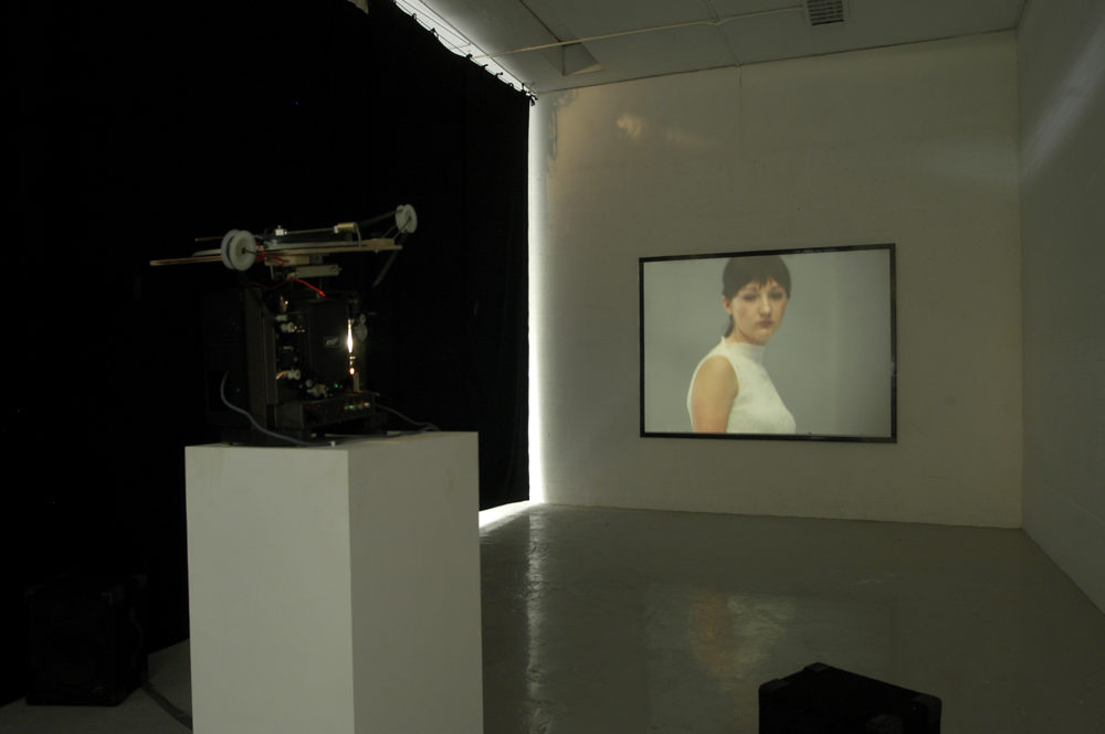 Installation view of Spin Cycle (2004). A projector screens a close up of a downcast looking woman on a wall.