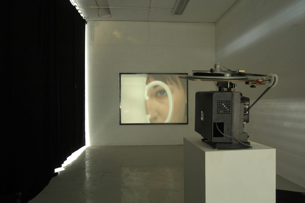 Installation view of Spin Cycle (2004). A projector screens a close up of a woman viewed through the handle of a jug on a wall.