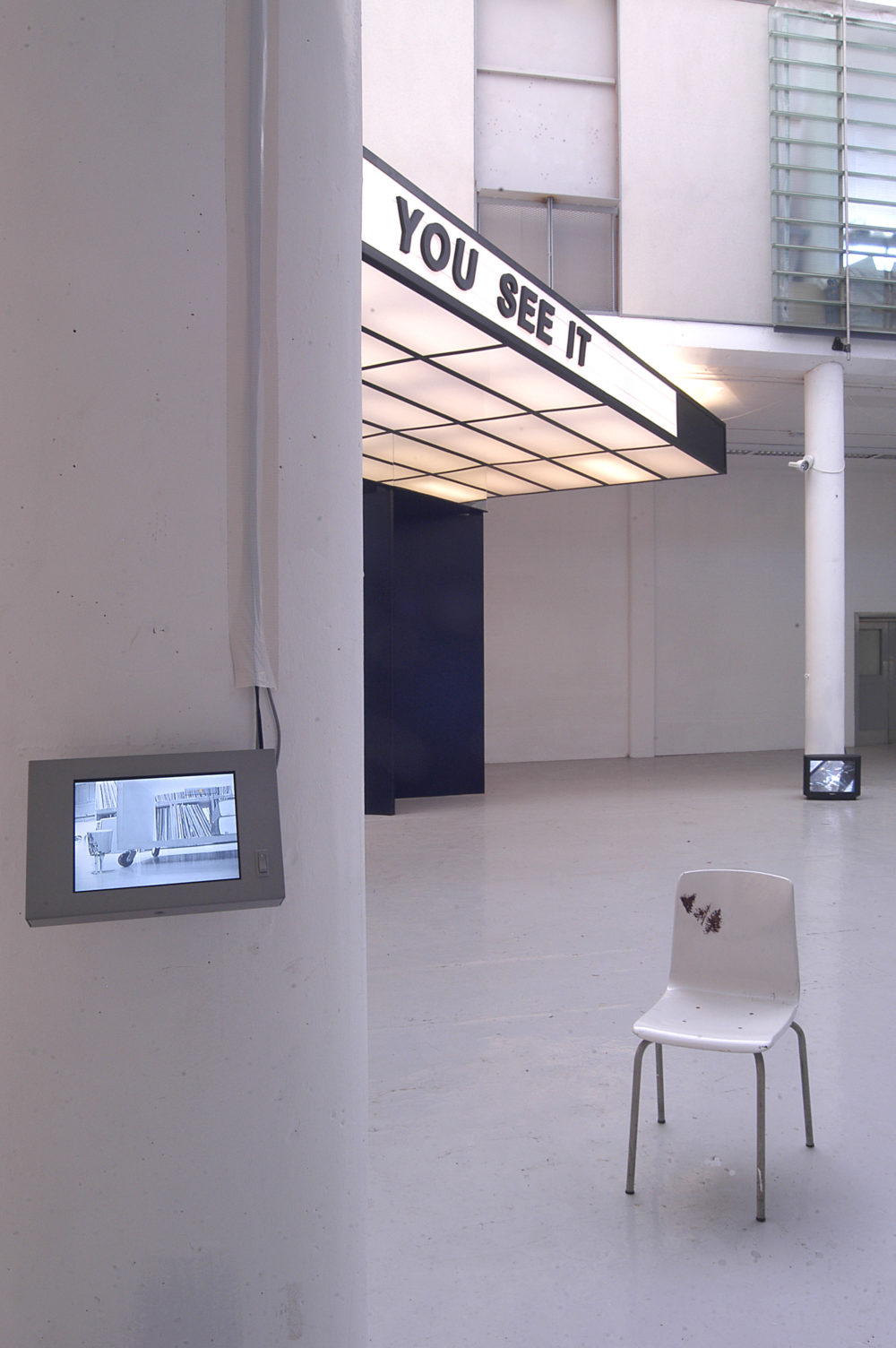 Installation view of Spin Cycle (2004). A small screen is attached to a pillar. Behind it is a plastic moulded chair.
