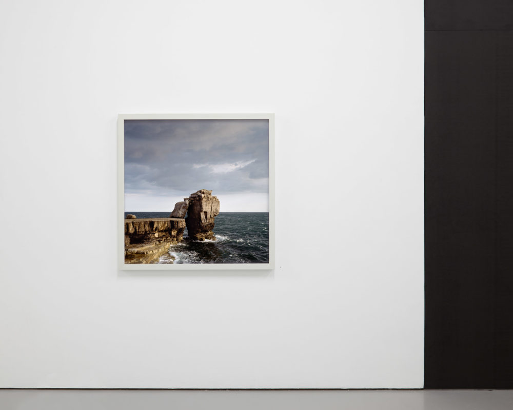 Install shot: A photo of crashing waves and a rocky outcrop is framed and hung on a white wall.