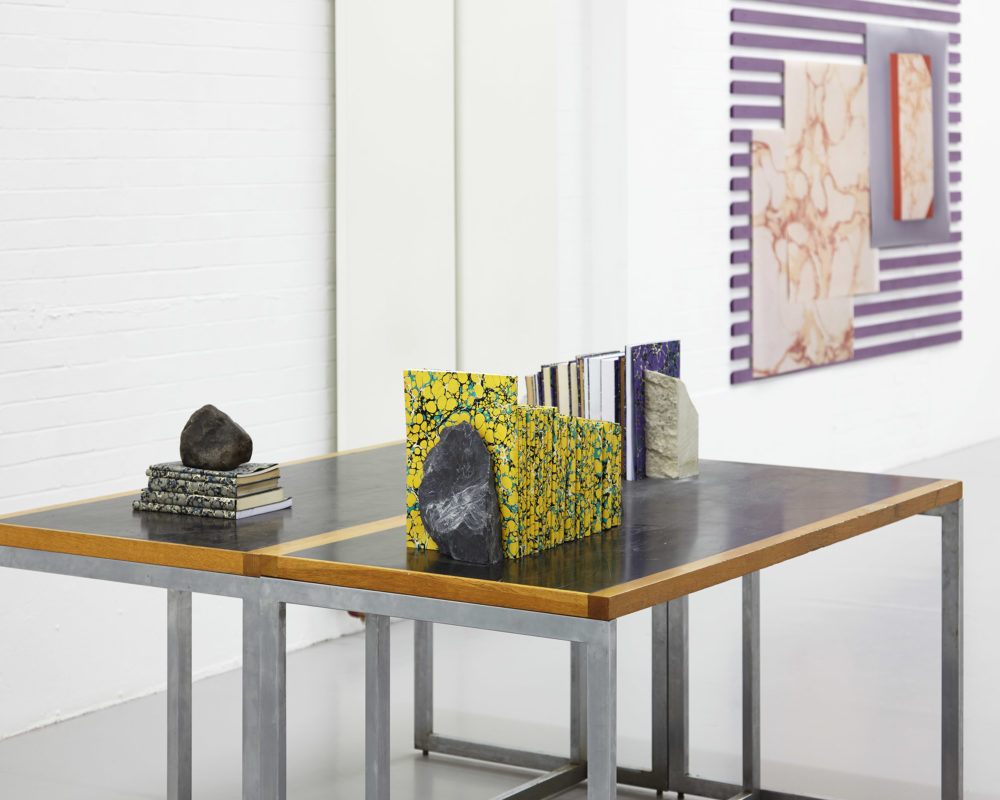 Installation shot: A table in the gallery has marbled paper covered books arranged atop it, using rocks as book ends.