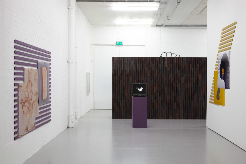Install shot: A screen stands on a plinth in front of a brick wall, to each side of the screen are corridor walls which show work made from printed photographs and slats of wood.