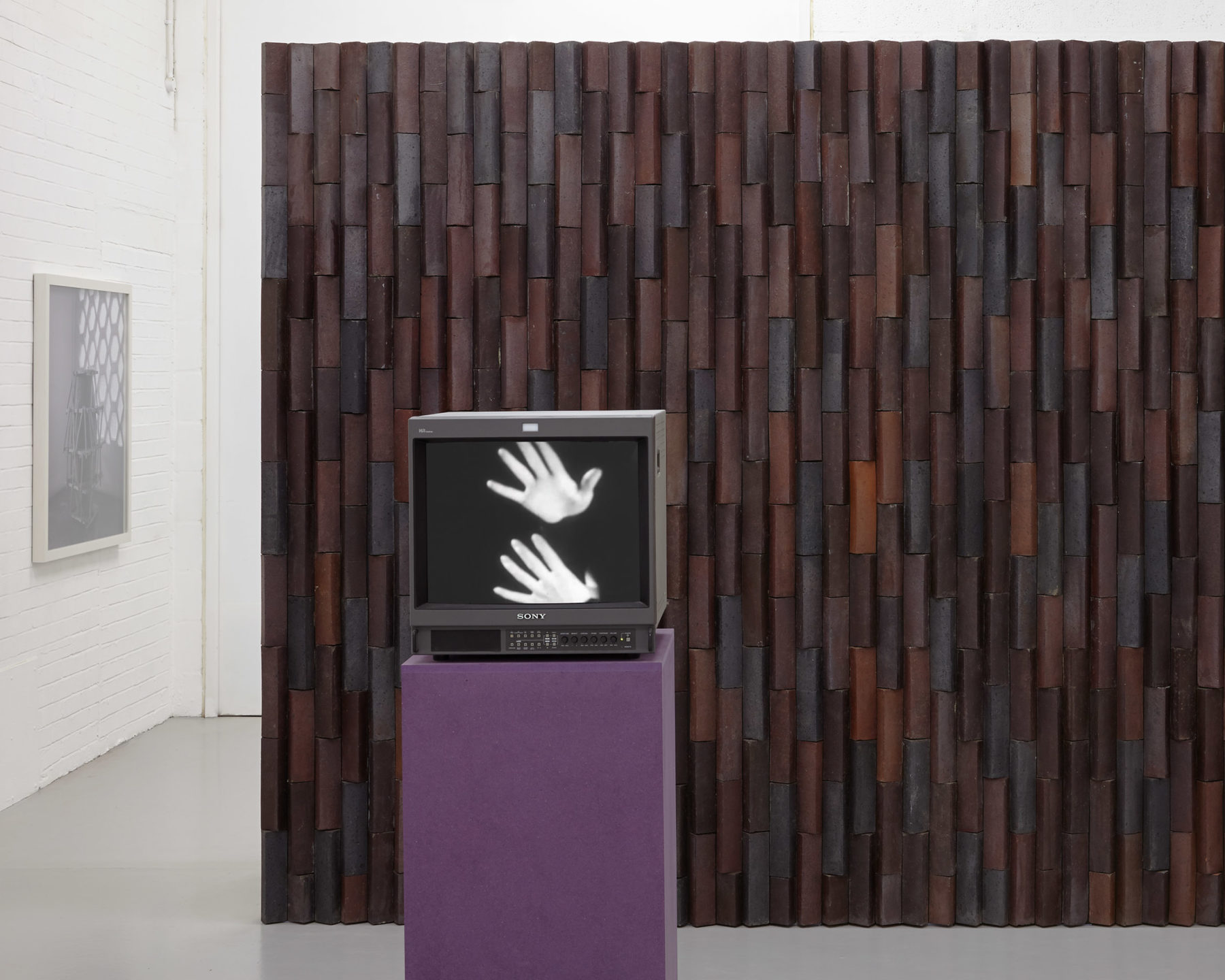 A screen in the gallery shows two white hands set against a dark background. This stands in front of a brick wall.