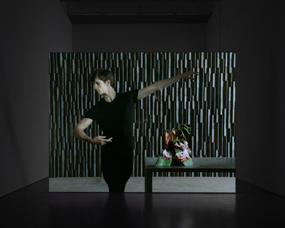Film still: A white person looks as though they are ballet dancing. Next to them is a table with an abstract sculpture on it.