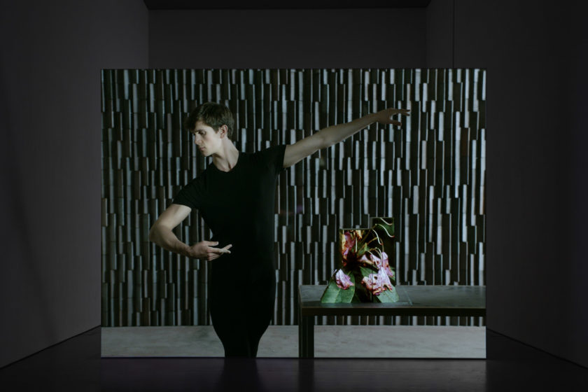 Film still: A white person looks as though they are ballet dancing. Next to them is a table with an abstract sculpture on it.