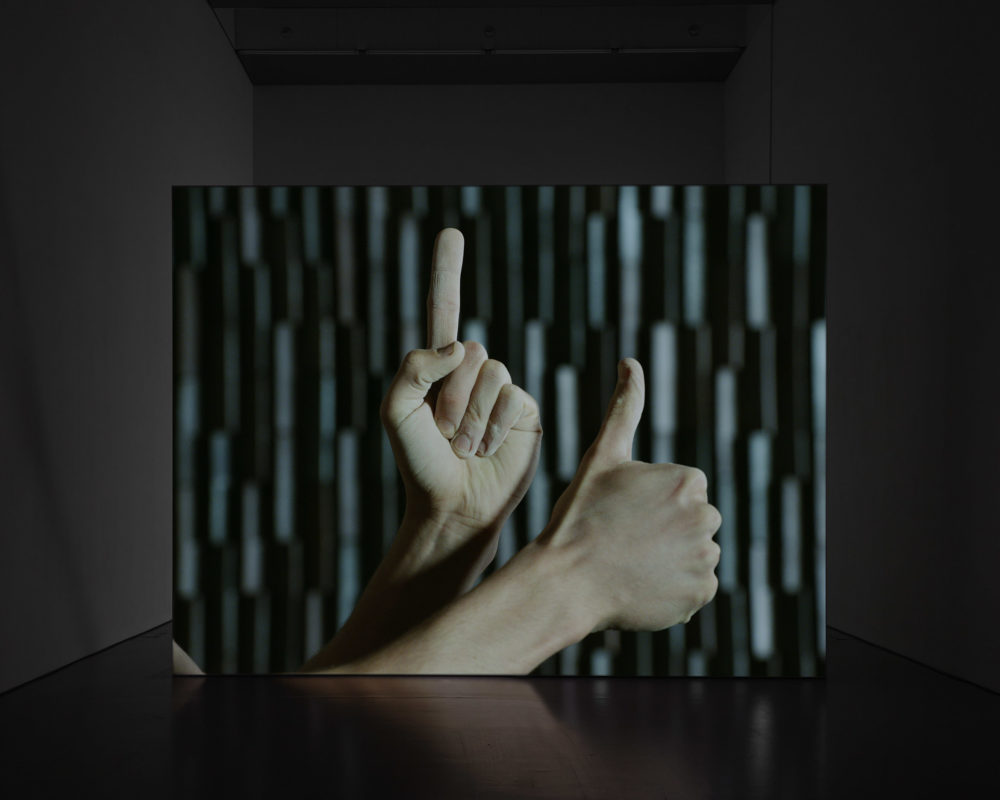 Film still: A white person holds up their hands, all that is in shot is their hands and a background. One hand has an extended index finger, the other has an extended thumb.