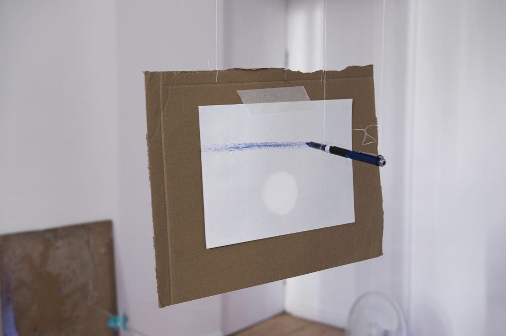 A blue pen is suspended from the ceiling horizontally. A white piece of paper is also suspended, but vertically. The blue pen is drawing an uneven line on the paper.