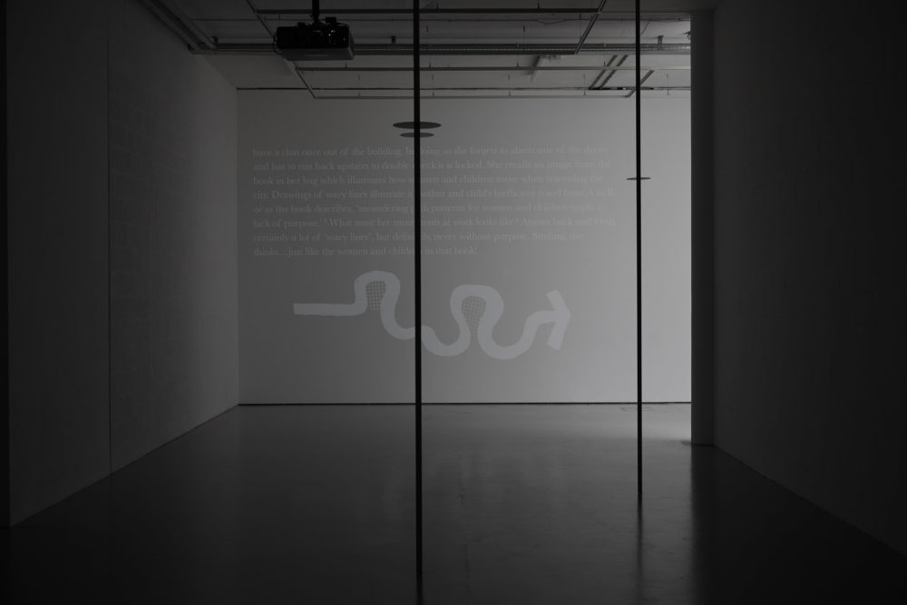 A corner of the gallery is dimly lit with two poles stretching from the ceiling to the floor in the middle of the walkway. The farthest wall has a projection on it of some text and a winding arrow pointing right.