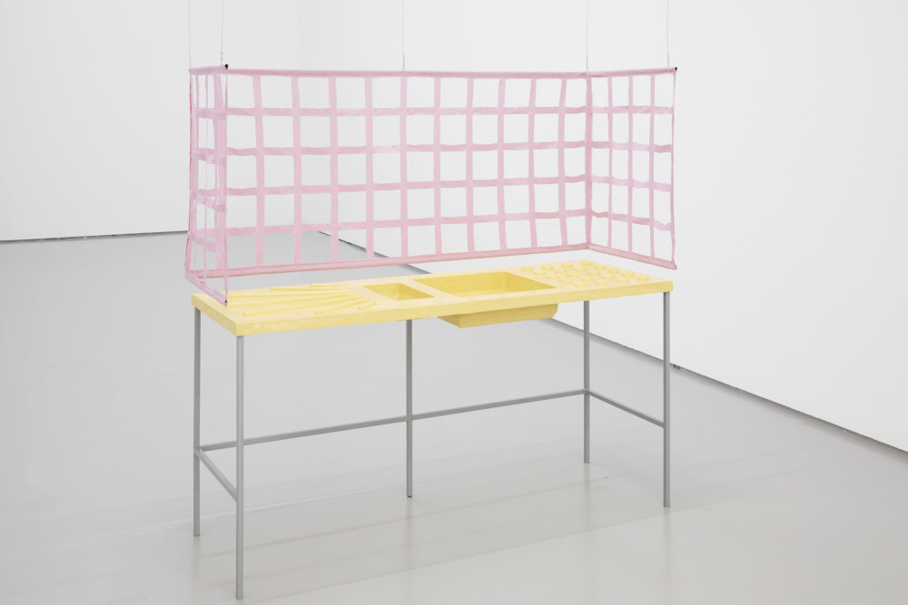 A kitchen sink has been rendered from fibreglass, painted yellow and displayed in a white gallery. There are no taps. There is a pink fabric grid hanging above and around it, evoking walls around a sink.