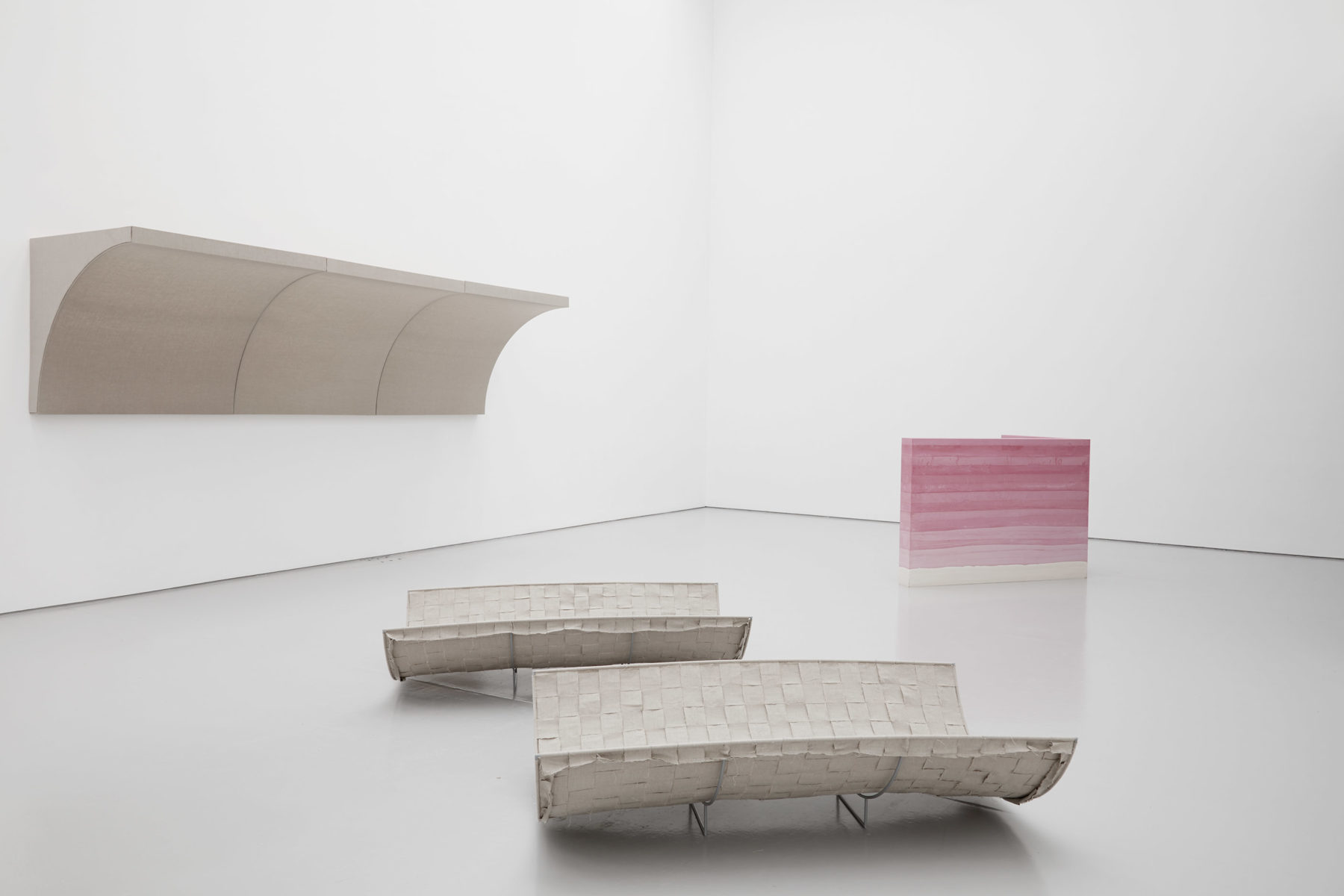 Bed like sculptures sit in the foreground of the gallery. Behind them is an overhanging cove like shelf and a pink striped wall.