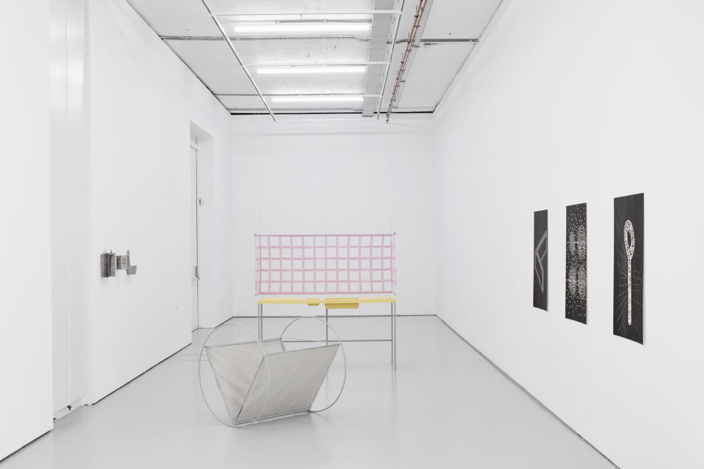 A kitchen sink has been rendered from fibreglass, painted yellow and displayed in a white gallery. In front of it is a structure that looks as though it rocks on large metal circles. The walls have large black and white prints.