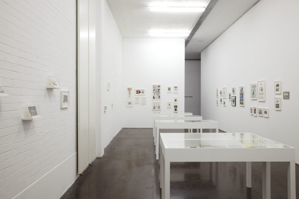 Installation view of The Artists' Postcard Show (2012). Five white table plinths with perspex tops hold postcards, as do frames on the wall.