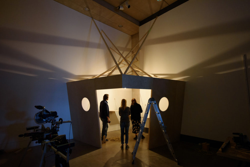 Installation shot: Three technicians work to erect a wooden structure in a white walled gallery.