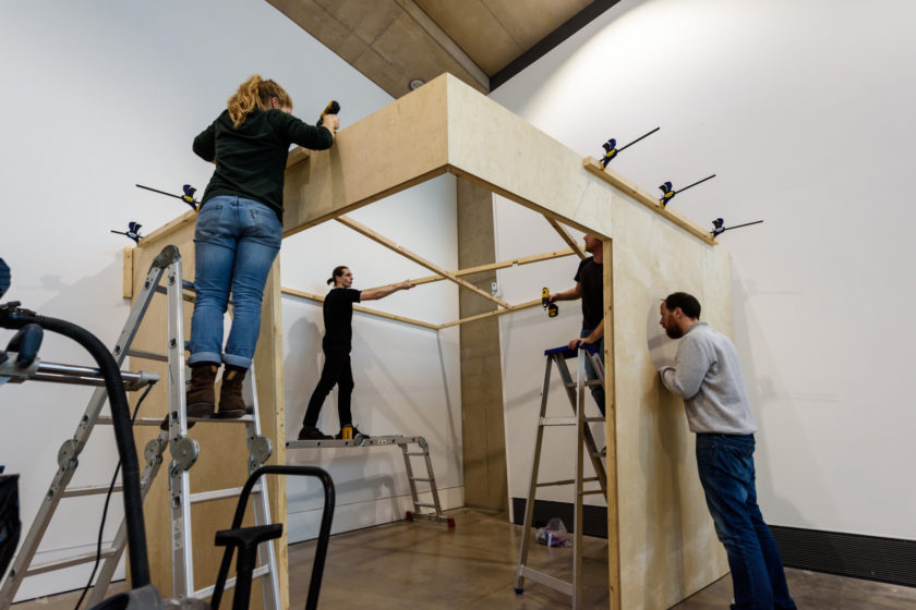 Installation shot: Four technicians work to erect a wooden structure in a white walled gallery.