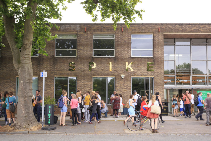 Photograph of the spike island building, a long flat roofed brick building with large windows and the metal letters S P I K E are on the front of the building. A lot of people gather outside chatting, on a summers day.