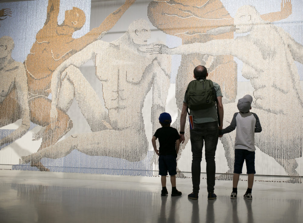 Three visitors stand in front of a large bead curtain decorated with large, nude figures of differing skin tones. The visitors look as though they're a family - father and sons. The sons stand either side of the father and hold his hand. They all study the curtain closely.