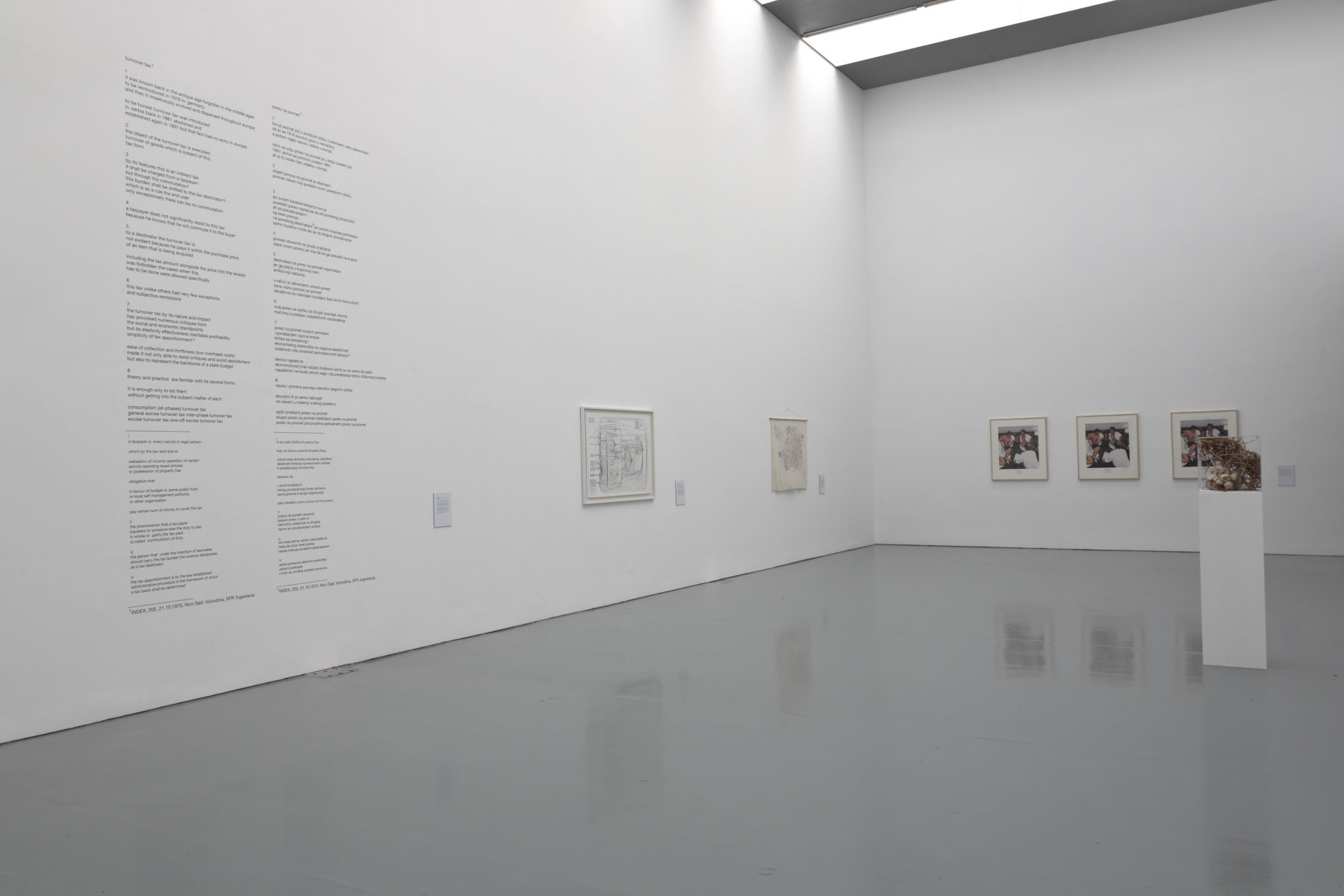 Installation view of Wealth of Nations (2010). The gallery is light and the walls are white. Five pictures hang on the walls and a plinth has a sculpture made from twigs atop it.