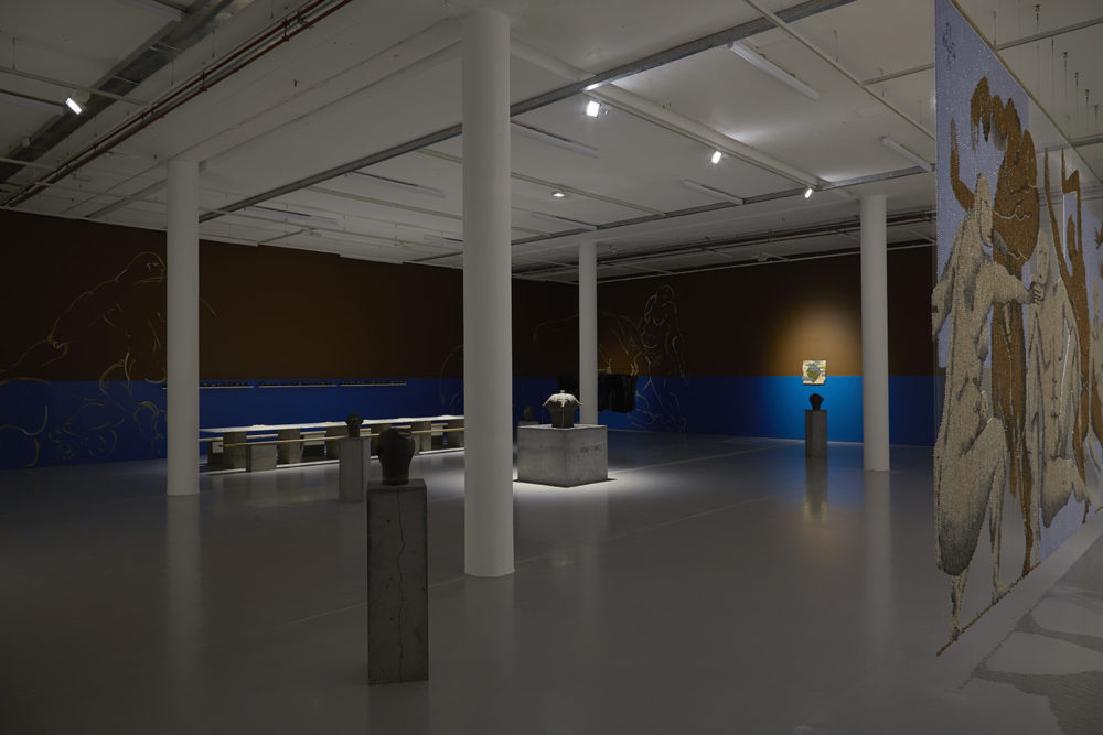 In the centre of the gallery lies a large cuboid concrete shape with a head-shaped item in the middle. There are four plinths with head-shaped things on top. A large bead curtain hangs from the ceiling, the beads illustrating a scene of lots of large nudes.