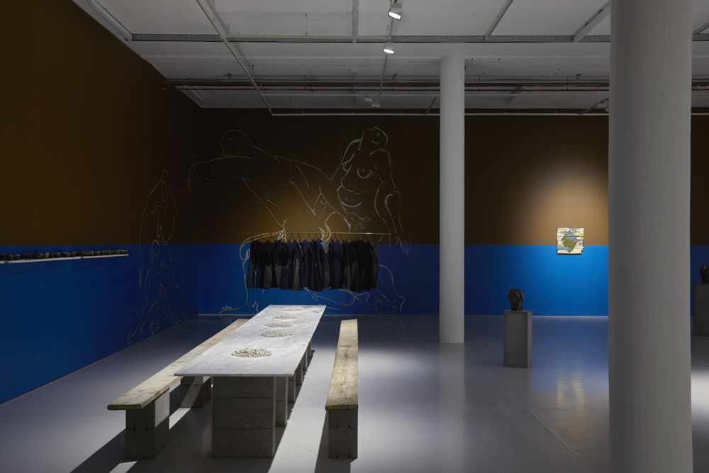 A large, long table runs the length of one of the walls of the gallery. It has two long benches either side, too. You can also see a plinth with a head shaped item on it.