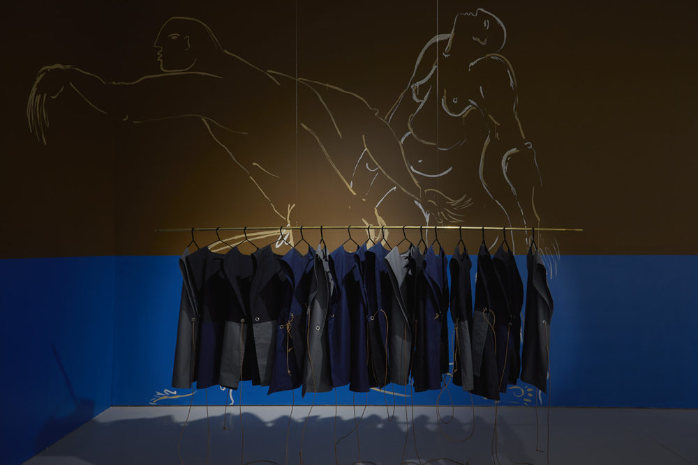 A rail hangs from the gallery ceiling, and hanging from that rail are sixteen utility aprons made from canvas and denim. Behind the rail, on the wall, are two large nude painted figures.