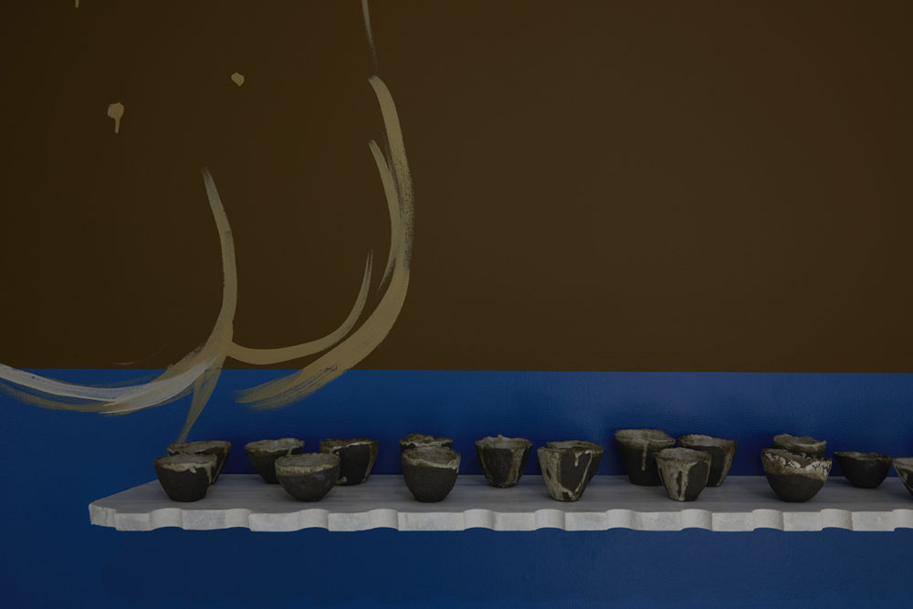 A white shelf with lots of rustic ceramic cups on top is attached to the gallery wall, on the blue bit. On the wall also is a painting of a large nude - all that is in this shot is the nude's bottom.