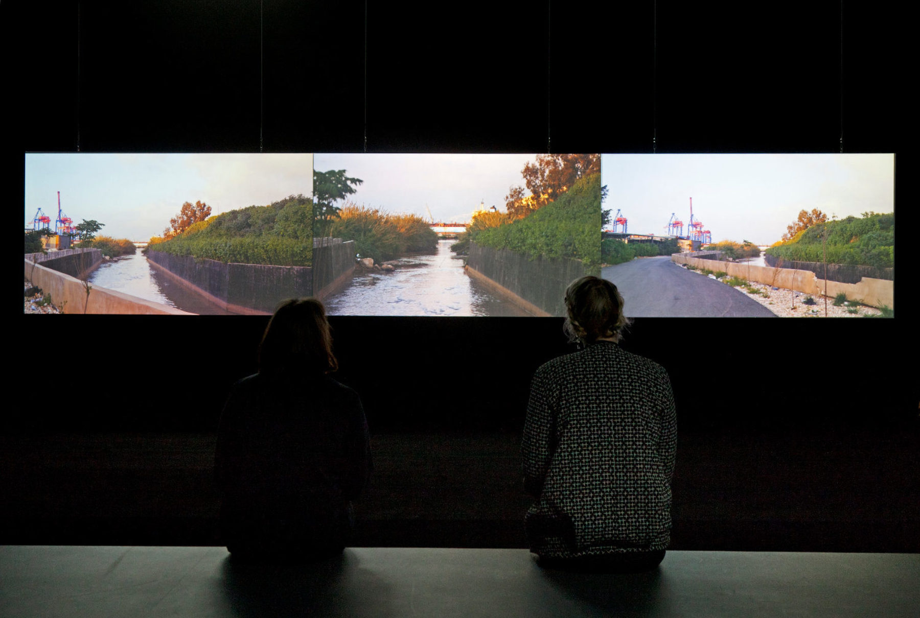 Installation shot: Two visitors study a screen showing three shots of a river.